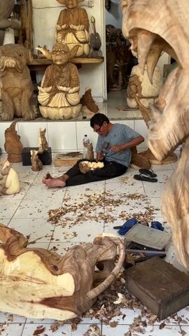 A medium shot shows a man sitting cross legged on a white tile floor and carving a small wooden animal figurine with hand tools, surrounded by larger wooden sculptures and piles of wood shavings. The man is wearing glasses and a blue t-shirt and dark pants, and is barefoot.  Behind him on a shelf are several Buddha-like sculptures. In the foreground is a large, partially carved bear sculpture with gaping jaws. A water bottle sits to the man's left. The scene conveys a cluttered workshop environment focused on wood carving.