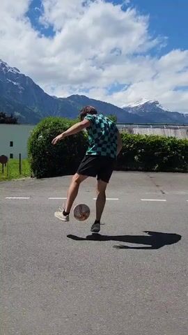 A man in a checkered shirt and black shorts is juggling a soccer ball with his feet and knees on an asphalt road. He is facing away from the camera and towards a backdrop of green trees and mountains. The mountains are partially covered in snow. The sky is blue with some clouds. There are a few buildings visible in the background, as well as a parked black car. The man is standing between two white lines painted on the road. His shadow is cast on the asphalt in front of him. The lighting is bright and sunny.