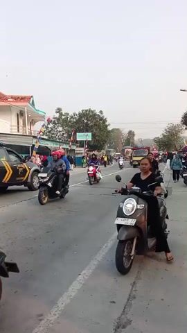 The video begins by showing a group of people riding motorcycles and cars on a road. There is a police car in the middle of the road, and several other vehicles are moving in the same direction. The people are mostly dressed in casual clothes. The road appears to be a rural or suburban area, with buildings and trees visible on the sides of the road. The sky is overcast, and the lighting suggests it is either early morning or late afternoon. There are flags and decorations on the buildings, indicating some sort of event or celebration is taking place. The video ends with a shot of a truck driving down the road.