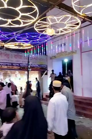 The video captures a bustling outdoor gathering at night, likely a market, fair, or festival, in Charfasson, Bangladesh. Numerous people, including men, women, and children, are seen moving through the crowded space.

The scene is brightly illuminated by extensive decorative lighting. Overhead, large circular and star-shaped patterns of string lights in yellow, blue, green, and red hues create a festive atmosphere. Additional vertical string lights and a prominent white light decoration shaped like a stylized rose are also visible. Stalls or temporary structures, also brightly lit, line the background.

People are actively walking, browsing, and socializing. Men wear a mix of casual attire like t-shirts and shirts, with some also wearing traditional prayer caps (kufi). Women are dressed in various styles, some opting for hijabs. A child is seen being carried by an adult. The ground appears to be unpaved, possibly dirt or sand.

In the background, a banner with text, likely in Bengali script, is visible but too distant and blurry to be clearly legible. The overall mood is lively and engaging, with people enjoying the vibrant evening event.