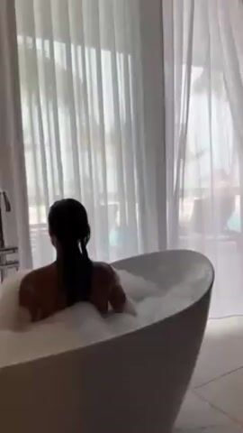A woman is sitting in a bubble bath, facing away from the camera, then turns her head slightly to the left. She has dark, wet hair pulled back into a low ponytail. The bathtub is a modern, egg-shaped design, filled with white bubbles. Behind the tub is a large window covered with sheer white curtains, allowing some light to filter through. The room has light-colored tile flooring and a chrome faucet is visible next to the tub.