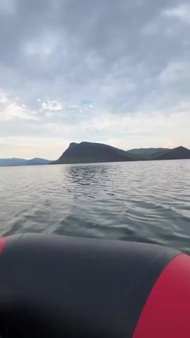 A low-angle, handheld shot shows the edge of an inflatable boat, likely red and black, moving through calm, slightly choppy water. In the distance, a large, dark, mountainous landmass dominates the horizon under a cloudy sky. The clouds are a mix of white and dark gray, with some lighter patches suggesting breaks in the overcast. The overall impression is of a serene, natural landscape, possibly a lake or large body of water, viewed from a recreational craft.