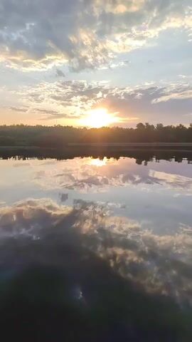 The video captures a serene sunset scene over a calm body of water, possibly a lake or river. The sun is setting on the horizon, its light partially obscured by a layer of clouds, creating a warm and radiant glow. The sky above is filled with a mix of clouds, some darker and some lighter, contributing to the overall atmospheric effect. 

The water surface acts as a natural mirror, reflecting the colors and patterns of the sky and clouds, enhancing the visual symmetry of the scene. A silhouette of trees and vegetation can be seen along the opposite bank, providing a dark contrast to the bright sky and water. There is no human presence or activity observed in the video. The scene evokes a sense of tranquility and natural beauty.