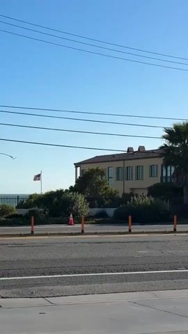 A gray pickup truck drives past a large tan house with a blue sky overhead. An American flag waves on a pole in front of the house. The road is paved, and there are orange traffic cones and lush green bushes along the side. A palm tree is visible to the right of the house.