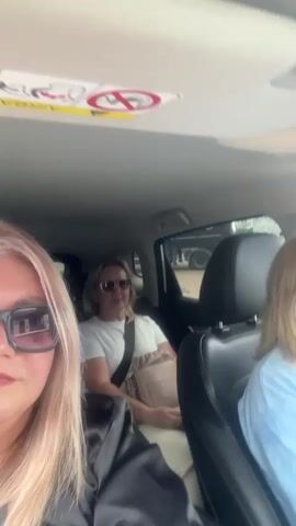 Three women are in a vehicle. The woman in the foreground has long blonde hair and is wearing sunglasses. She is looking at the camera. The woman in the middle has long blonde hair, is wearing sunglasses, and is wearing a white shirt with a tan cross-body bag. She is dancing in her seat. The woman in the background has long blonde hair and is wearing a light blue button-down shirt and pink sunglasses. She is smiling.