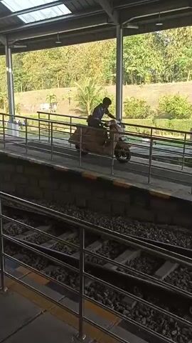 A person rides a scooter with a cardboard-covered body down a ramp at a train station. The scooter is brown and has a brown seat. The person is wearing a dark shirt and dark pants. The train station has a metal roof and a concrete platform. The train tracks are visible in the foreground. The background shows a grassy hill with trees. The sun is shining and the sky is blue.