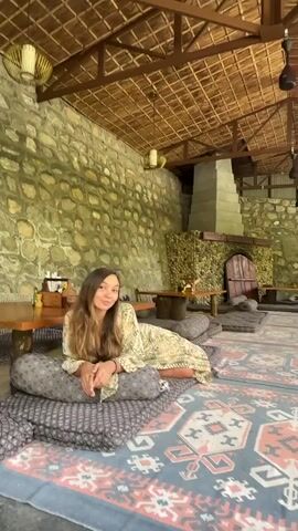 The video shows a woman sitting on cushions in an open-air room. The room has stone walls, a thatched roof, hanging lamps, and a large decorative fireplace. The woman is wearing a patterned robe and sits cross-legged on the cushions. She looks at the camera and smiles. The floor is covered with a large patterned rug, and there are low wooden tables in the room. The woman then turns to her left and looks around the room.