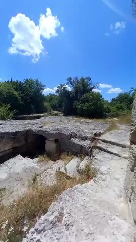 The video shows a rocky, overgrown landscape under a bright blue sky with scattered clouds. The foreground is dominated by rough, light-colored rock formations, some of which appear to be man-made structures with cavities and cut-out areas. Sparse dry grass and some small wildflowers grow among the rocks. In the middle ground, more dense green trees and bushes form a backdrop. The background reveals a vast, green valley stretching out to the horizon, with a clear demarcation between the elevated rocky terrain and the lower-lying landscape. The camera pans slowly, capturing the expansive view and the weathered texture of the stone structures. No people or animals are visible. The overall impression is one of ancient ruins or geological formations in a natural, sun-drenched environment.