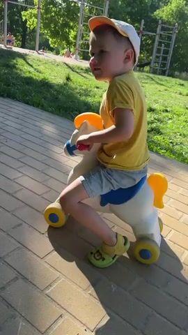 A toddler wearing a yellow shirt, denim shorts, green sandals, and a white cap rides a white plastic rocking horse on a brick-paved pathway in a park. The rocking horse has a blue seat and yellow accents. The toddler looks back at the camera while riding. Green grass, trees, and a playground structure are visible in the background. The light suggests it's daytime.
