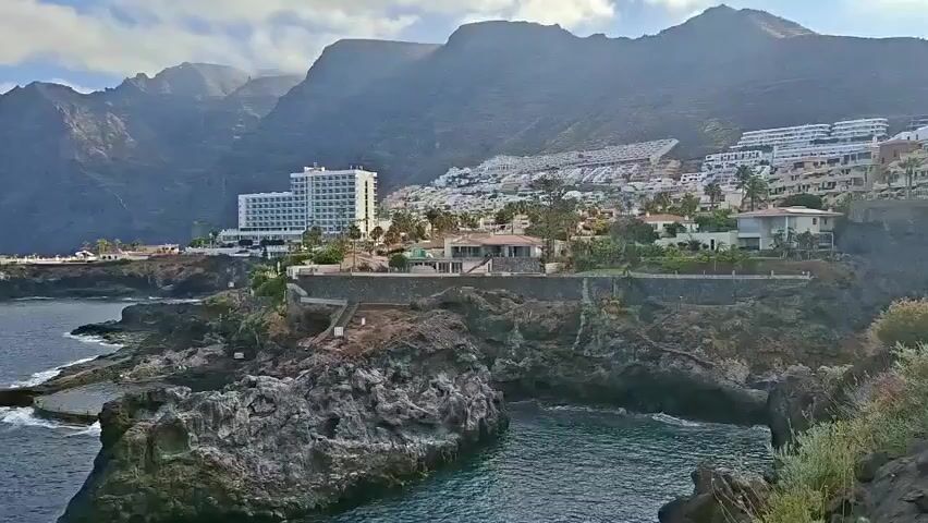This is a wide shot of a coastal area with rocky cliffs and the ocean. In the background, there are mountains and a town with white buildings, including a large hotel. The sky is partly cloudy. The water is dark blue, and there are some waves crashing against the rocks. There are no people or visible objects in the water. The scene appears to be a tourist resort.