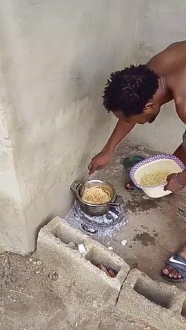 A man with dreadlocks, wearing shorts and sandals, stands in an outdoor cooking area against a light-colored wall. He is stirring cooked spaghetti or noodles in a silver pot using a spoon in his right hand. The pot sits on a charcoal stove made from cement blocks and charcoal. In his left hand, he holds a white colander filled with more cooked spaghetti or noodles. The ground is dirt, with several cement blocks and small pieces of debris visible.