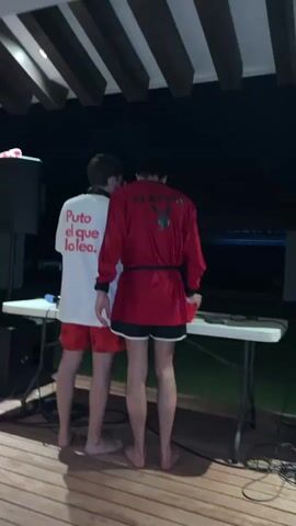 Two shirtless men stand outdoors, seemingly at night, on a wooden deck under a slatted roof. A table in front of them holds DJ equipment, connected with several visible cords. The man on the left wears a white t-shirt with red text, "Puto el que lo lea," and red patterned shorts. The man on the right wears a red long-sleeve shirt with "PLAYBOY" on the back, a black robe tie, and black shorts. A speaker is positioned near the left edge of the table. Music plays in the background. One of the men holds what appears to be a cigarette.