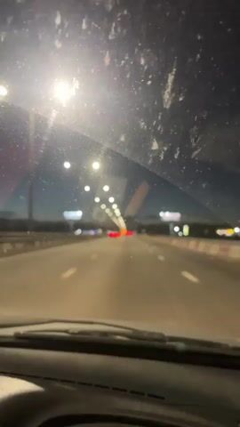 The video shows a first-person perspective from inside a car driving on a highway at night. The windshield is dirty and smudged, with visible dirt and streaks. Streetlights line the highway, casting a bright glow and creating lens flares. In the distance, the red taillights of other vehicles are visible. The sky is dark blue, indicating it is either early evening or late night. The dashboard and part of the steering wheel are visible at the bottom of the frame. A Russian flag is partially visible on the left side of the dashboard. The overall impression is a journey on a dimly lit highway during nighttime.