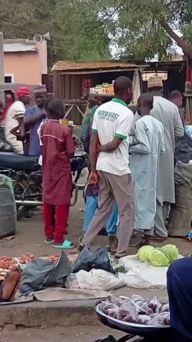 The scene shows a bustling outdoor marketplace. People in various attire, including traditional clothing, are moving about. Some individuals are gathered around vendors, examining goods like vegetables. A person wearing a shirt that reads "World Roast" stands out in the crowd. The camera shifts perspective, showing different angles of the market and the diverse array of people present. The atmosphere suggests a lively commercial exchange.