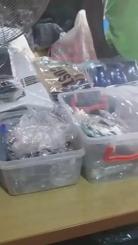 The video shows a close-up of a table covered with various items, possibly in a market or a shop. In the foreground, there are two clear plastic containers with orange handles, filled with small, shiny objects that appear to be hardware or accessories. To the left of these containers, there are more items packaged in plastic, including what looks like fabric or clothing. Behind these containers, more packaged goods are visible, including some with blue packaging and one with a label that says "SUN". A person wearing a green t-shirt is partially visible in the background, standing behind the table. The lighting is dim, and the overall impression is of a busy retail environment.