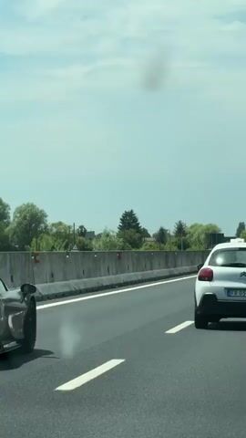 The video shows two cars on a highway. The car in the foreground is black and appears to be a sports car, with white smoke coming from its rear. The car in the background is white and is following behind the black car. Both cars are driving in the right lane of the highway. The sky is cloudy, and there are trees and a concrete barrier visible on the side of the road.