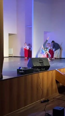 On a stage, several young girls in traditional red and white costumes stand to the side as a man in a dark suit and white pants directs two other girls in modern dance attire. The two girls are performing a choreographed dance, while the other girls and the man watch. The stage is lit with a blue hue and has a wood-paneled front. A large black speaker sits prominently at the front of the stage, and wooden stairs are visible on the right side. The environment suggests a school performance or talent show.
