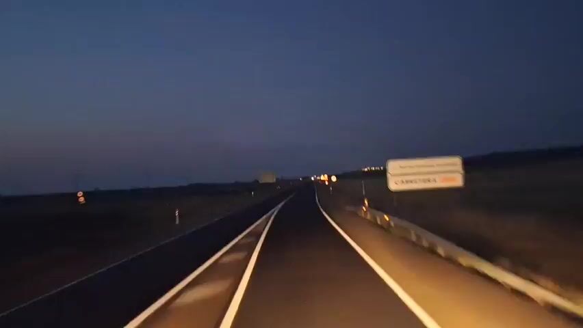 The video captures a perspective from inside a vehicle at dusk or dawn. The road ahead is visible, with white lines marking the lanes. The sky displays a gradient of dark blue to orange, indicating low light conditions. On the right side of the road, a guardrail and a sign are partially visible. In the distance, there are some lights and what appears to be a transmission tower. The surrounding landscape is flat and appears to be open country. The video is slightly shaky, suggesting movement from the vehicle.