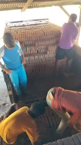 Four individuals are shown in a brick yard, some are carrying or stacking bricks. The environment appears to be an open-air structure with a wooden roof. The people are wearing casual clothing. One person is wearing a blue tracksuit, another in a yellow t-shirt, another in an orange t-shirt, and one in a purple t-shirt. The bricks are stacked in large piles. The lighting suggests it is daytime.