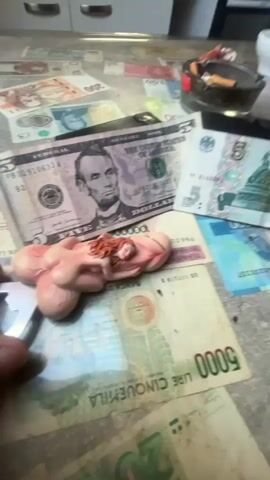 The video shows a close-up of a cluttered table surface covered with various foreign banknotes, an ashtray with cigarette butts, and a phallic-shaped figurine with a woman's head at the tip. The camera slowly pans across the table, focusing on the figurine and the money. The environment appears to be indoors, possibly a kitchen or dining area, with white cabinets and a stovetop visible in the background. The lighting is moderate, and the overall scene is somewhat grimy, with some dirt and smudges on the table. The figurine is the central focus, and its placement on top of the money suggests a symbolic or provocative intent. No people are visible in the video, only a hand briefly enters the frame to adjust the figurine.
