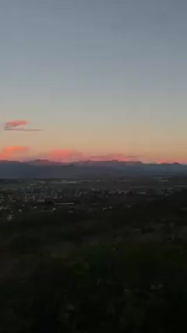 The video shows a wide shot of a landscape at dusk. A city, or large town, is visible in the valley below, with rows of buildings stretching across the scene. Behind the city, a mountain range rises, its peaks silhouetted against the fading light. The sky is a gradient of colors, from a deep blue higher up to a soft pink and orange near the horizon, where clouds are tinged with the colors of the sunset. In the foreground, dark green foliage partially obscures the view, suggesting the shot is taken from an elevated position. The overall scene conveys a sense of calm and scenic beauty.