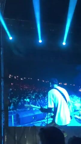 The video shows a large screen at a concert. The screen displays a man wearing a white t-shirt, dark pants, and a dark flat cap singing into a microphone. He is performing on a stage in front of a large crowd, which is visible in the background. Blue spotlights shine down from above the screen. The camera is handheld and slightly shaky. The perspective suggests the video was taken by someone in the audience.