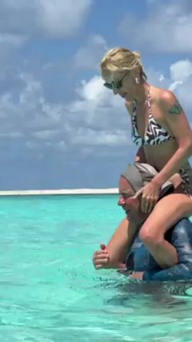 In a bright, outdoor shot under a sky of scattered clouds, a woman in a bikini sits on a man's shoulders in shallow, turquoise water. She wears a black and white patterned bikini and sunglasses, her blonde hair pulled back. The man is wearing a blue rash guard and a baseball cap. He's submerged up to his chest, gripping her legs for support. They are in the middle of clear, tropical water, with a white, sandy horizon in the background. The woman appears to be adjusting the man's cap, and they both seem to be enjoying the sunny weather and picturesque surroundings.