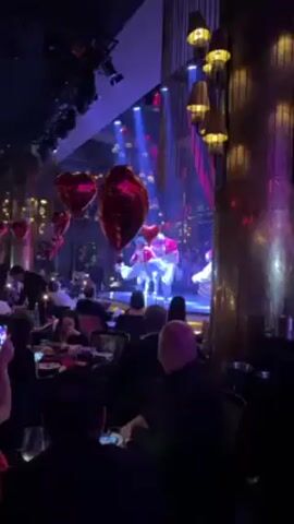 The video shows a group of dancers performing on a stage in what appears to be a restaurant or nightclub setting. The dancers, three in number, are dressed in pink and white outfits. They are executing a synchronized routine under bright stage lights, including blue and purple hues. The performance is happening in front of a seated audience who are eating and watching. Red heart-shaped balloons decorate the space, adding a festive or celebratory atmosphere. The room has ornate decorative elements, including gold-colored pillars and hanging light fixtures. The camera view is from the audience, slightly obstructed by people in the foreground.