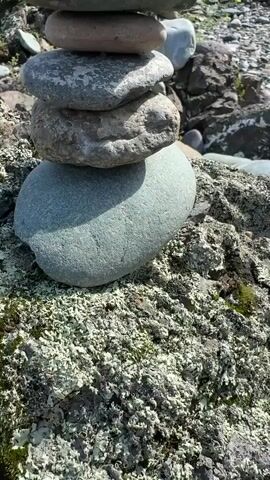 A video shows a stone cairn in focus, with other stone cairns and rocks in the background. The cairn in focus is made of stacked grey and greenish rocks. The background features a rocky, moss-covered terrain with sparse green vegetation. The lighting suggests it is daytime.