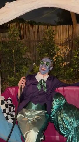 The video features a person in an elaborate costume, sitting on a bright pink couch in an outdoor setting at night. The individual's face is covered in blue-grey makeup, with white teeth painted on to create a wide, exaggerated smile. They are wearing small, round, dark sunglasses. Their attire includes a purple jacket, a white collared shirt, a prominent green bow tie, and shiny light green or silver pants with a dark green sash.

The person is holding a black hookah hose to their mouth, maintaining the wide-mouthed, laughing expression throughout the short clip. To their left on the couch is a black and white checkered cushion. To their right, a mass of shiny, dark green fabric is draped, possibly part of another costume or decoration.

The background consists of a tall wooden fence or bamboo screen, behind which lush green foliage is visible. The dark sky indicates it is nighttime, with artificial lighting illuminating the scene. The overall ambiance suggests a lively event or party, with the costumed individual posing in a theatrical manner.