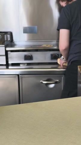 A person in a dark t-shirt and black pants stands in a kitchen area, facing away from the camera. They are near a stainless steel flat-top grill and some stainless steel cabinets. The person appears to be preparing food on the grill, although the specifics are not clear due to the angle and distance. Their left arm is visible, revealing a tattoo and a wristwatch. The background includes stainless steel surfaces, suggesting a commercial kitchen environment. A light-colored countertop is in the foreground.