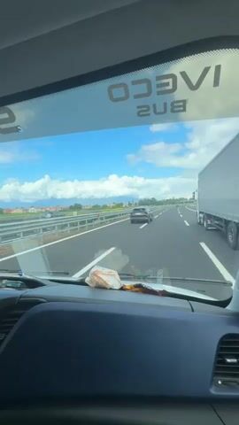 The video shows a view from inside a vehicle, likely a bus or truck, looking out through the windshield. The vehicle is traveling on a highway. The road is paved and has lane markings. There are guardrails along the side of the road. In the distance, there are fields, trees, and buildings, suggesting a rural or suburban environment. The sky is blue with white clouds. There is a white vehicle ahead on the highway. The dashboard of the vehicle is visible in the foreground, with some items on it. The word "IVECO BUS" is visible, reversed, on the top of the windshield, indicating the type of vehicle. The audio is of a person speaking in Romanian, but the content of the speech is not relevant to the visual description.
