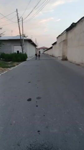 The video captures a street scene in Andijan, Uzbekistan, likely during the late afternoon or early evening due to the soft, diffused light and the pale sky with hints of orange and blue. Three individuals, appearing to be children or young adolescents, are walking down a paved road. Two of them are closer together and appear to be interacting or walking side-by-side, while the third, slightly ahead, walks alone. They are all dressed in casual clothing, typical for warm weather.

The setting is residential, with buildings lining both sides of the street. The structures on the left are lower and appear to be single-story homes or outbuildings with some vegetation in front. On the right, a long, tall, cream-colored wall dominates the view, suggesting a property boundary or the side of a larger structure. Overhead, utility wires are visible, crisscrossing the sky, a common sight in many urban and semi-urban environments. The road itself is asphalt and shows signs of wear, with dark patches that could be oil stains or damp spots.

The overall mood is peaceful and ordinary, depicting a simple moment of children walking in their neighborhood. There is no audible sound, so any emotional interpretation is based solely on the visual cues. The children's posture and gait do not suggest any particular urgency or distress. The absence of strong shadows indicates the sun is not directly overhead, supporting the idea of it being late in the day. No text is visible in the video.