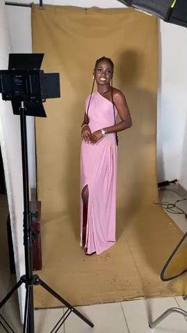 A young Black woman stands facing the camera in a photography studio. She is wearing a long, pink, one-shoulder dress with a thigh-high slit and gold embellishments on the hip. Her hair is styled in braids. She is standing in front of a tan backdrop. To her left is a large LED light on a stand. To her right, partially visible, is another light fixture with an umbrella. The floor is covered with a tan tarp. The video pans slightly to the right, showing more studio equipment and a shelf with decorative items. The video then shifts focus to a person holding a camera, with parts of other people and studio elements in the background.