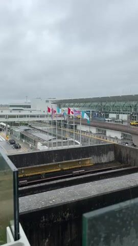 The video shows an airport terminal on an overcast day. Flags of various nations are displayed on poles in front of the terminal. Cars and a train are visible on the airport grounds. The scene appears to be a typical airport environment with travelers and staff moving about. The weather is cloudy, suggesting it might be morning or late afternoon. The location is Richmond, Canada. There is no visible text in the video.