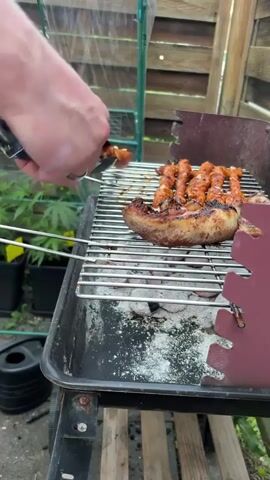 A hand holding tongs is shown flipping a large sausage on a grill. The sausage is partially cooked and appears to have grill marks. To the right of the large sausage, there are several smaller sausages arranged in a row. The grill contains hot coals. The background shows a greenhouse and wooden fencing. The scene appears to be an outdoor setting.