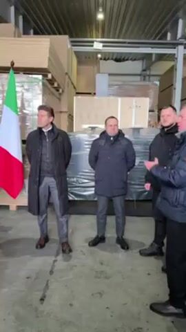 The video shows a medium shot of three men standing in a warehouse with the flags of the European Union and Italy behind them. The man in the middle wears a long black coat over a grey suit. The man to his right wears a navy blue jacket. A third man stands to the right, partially cut off by the frame. The warehouse appears to be filled with stacks of plywood and boxed items, and it is lit by overhead lights. Someone is speaking.