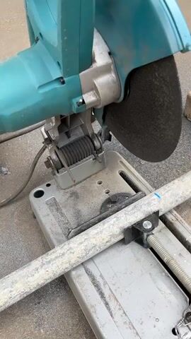 A metal chop saw is shown cutting a metal pipe. The saw has a teal-colored body and a black cutting wheel. The pipe is clamped to the saw's base. Sparks are flying from the cutting area. A gloved hand is operating the saw's handle. The environment appears to be outdoors, with a concrete or asphalt surface visible.