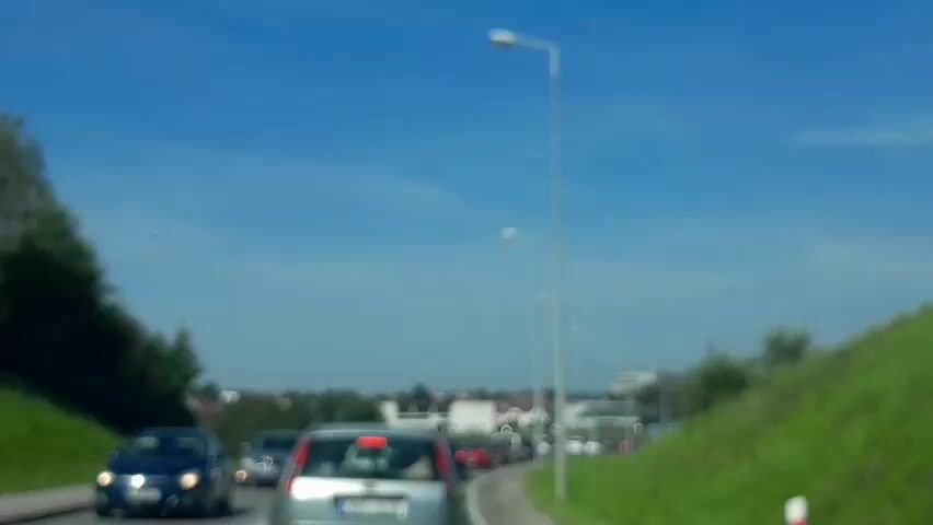 The video shows a view from a car in traffic on a sunny day. Several cars are visible on the road. On the right side of the road, there is a grassy embankment with trees and a lamppost. In the distance, there are buildings and a church steeple. A small orange object is visible in the sky at the top left of the frame. The text overlay indicates "Proviv vsye utro, synok," which translates to "I spent all morning, sonny."