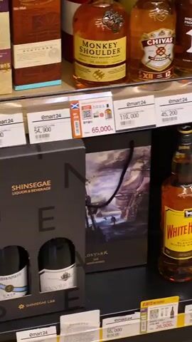 The video displays a close-up view of a liquor store shelf, likely a convenience store given the "Emart 24" branding. The shelves are stocked with various bottles of whiskey and other spirits. Several bottles of Monkey Shoulder, Chivas Regal, and Nikka are visible. There are also bottles of White Horse and a blue bottle labeled "Relax Riesling." Smaller bottles and gift sets are also present.

The shelves are organized with price tags prominently displayed, many showing prices in Korean Won, such as 66,500, 45,200, and 47,000. Some tags have QR codes, suggesting potential promotions or payment options. Packaging for gift items is also visible, including a dark box with a ship illustration and the text "LOSTARK."

The lighting suggests an indoor setting, likely during daylight hours given the brightness. The overall atmosphere is that of a retail environment focused on alcoholic beverages. The video pans across the shelves, providing a detailed look at the product selection and pricing. No people are visible in the footage. The prominent branding of "Emart 24" indicates the specific location within South Korea.