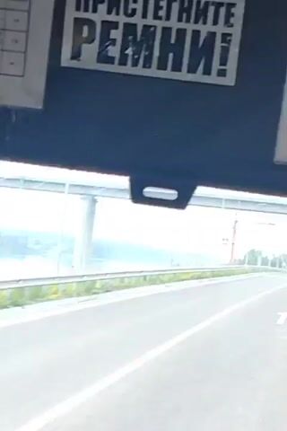 The video shows a view from inside a vehicle, possibly a bus, looking out a window. The foreground is dark, suggesting the interior of the vehicle. The middle ground shows a road with white lane markings and a guardrail along the side. Beyond the guardrail is a body of water, likely a river, with trees on the far bank and a bridge structure in the distance. The sky is overcast and bright. There are no people visible in the video. The vehicle appears to be moving.