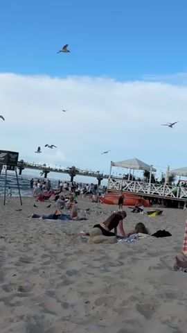 A beach scene captured from a low angle looking towards the water. The sand in the foreground is textured and scattered with a few small objects. In the middle ground, several people are relaxing on the sand. Two individuals in the immediate foreground are lying on their stomachs, with one facing away from the camera and the other lying on their side. Behind them, more people are spread out on towels and blankets. Further back, a pier extends into the water, crowded with people walking and standing. To the right of the pier, a white structure with a canopy and tables and chairs suggests a beachside establishment. An orange inflatable object, possibly a life raft, is visible near the structure. Seagulls are flying in the blue sky, which is dotted with white clouds. The overall atmosphere appears to be a busy, recreational day at the beach.