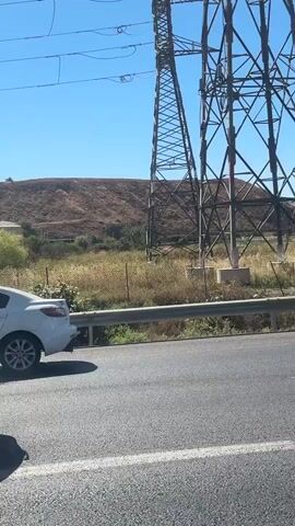 The video depicts a white sedan engulfed in flames on the side of a road during the daytime. Initially, smoke emanates from the vehicle, which is parked next to a metal guardrail. Large power pylons and overhead power lines are visible in the background, set against a clear blue sky and distant hills. As the video progresses, the fire intensifies, producing thick plumes of black smoke that rise into the sky. Other vehicles, including a white Honda Civic (license plate "62-42-31") and a white Volvo XC40 (license plate "476-49-703"), drive past the burning car on the multi-lane road. An emergency siren can be heard in the background. The scene is outdoors, indicating a rural or semi-rural setting with visible dry vegetation beyond the guardrail.