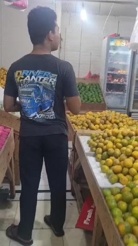 Here is a concise description of the video:

A young man is shopping for fruit at a fruit stand. He is wearing a black t-shirt and black pants. The shirt features a graphic design of a truck. The fruit stand displays rows of oranges and avocados. The man looks around and speaks briefly to someone off-camera. He is holding a plastic shopping bag in his hands. There is a refrigerator full of fruit behind him.