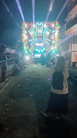 The video shows a dimly lit outdoor scene at night, possibly a street or an open area, with a large, elaborate sound system as the central focus. The sound system is mounted on a vehicle, possibly a truck, and is adorned with numerous flashing lights, predominantly in shades of blue, green, and white, creating a vibrant and dynamic visual display. Beams of white light emanate from the top of the sound system, illuminating the night sky. Several people are visible in the foreground and midground. In the foreground, a person wearing a headscarf and a backpack is standing, facing away from the camera and towards the sound system. To the left of the sound system, a group of people are seated or crouching on the ground, facing the spectacle. Others are standing around. The ground is littered with debris, suggesting a public gathering. The overall atmosphere appears to be that of an outdoor event or concert.