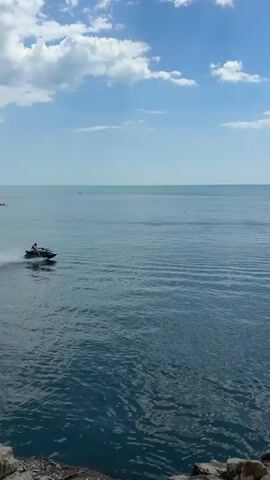 A jet ski is moving quickly across the water, creating a spray. A person is riding the jet ski. In the background, there are clouds in a blue sky and a distant sailboat. Another person is kayaking further out in the water. The water is blue and has ripples. Rocks are visible in the foreground.