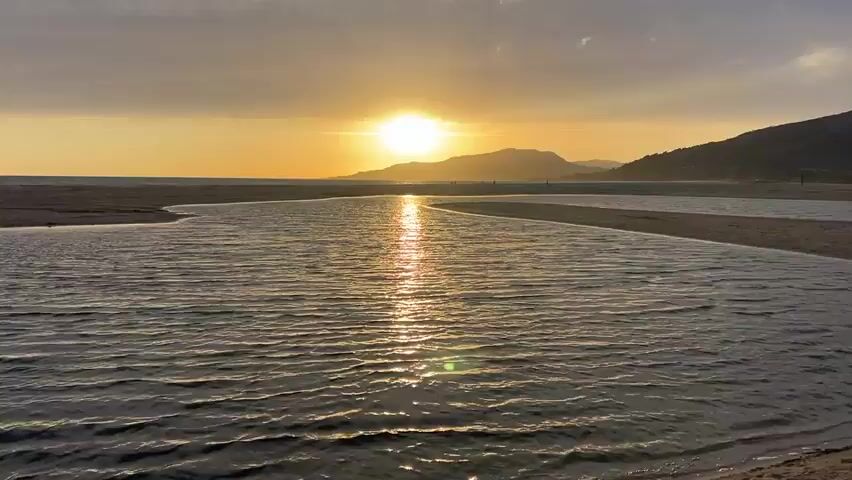 The video shows a sunset over a body of water. The sun is bright and positioned in the center of the frame, casting a golden glow on the scene. The water is calm with gentle ripples, and its surface reflects the sunlight. In the background, there are hills and a sandy shore. The sky is a mix of yellow and orange hues, with some clouds present. There are a few indistinct figures visible on the shore in the distance. The overall scene is peaceful and serene, capturing the beauty of nature at dusk.