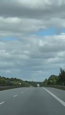 A forward-facing shot shows a multi-lane highway with trucks driving in the distance. The highway is bordered by lush green trees on both sides. The sky is cloudy with patches of blue. There is a significant amount of standing water on the left side of the highway, creating a reflection of the trees and sky. A white truck with a red stripe is visible in the distance on the left, and a white truck with a trailer is in the center of the frame on the right. The road markings, including solid white lines on the right edge and dashed white lines in the middle, are clearly visible. The overall impression is a wet highway scene under a partly cloudy sky.