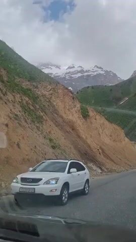 The video shows a road alongside a steep, bare, light brown hillside, with snow-capped mountains in the background under a cloudy sky. The road surface is gray asphalt. There is some sparse vegetation on the left side of the hillside and more lush green growth on the right. A patch of snow is also visible on the right side of the frame. The camera is positioned low, likely from inside a car. The scene conveys a sense of remote mountainous terrain.