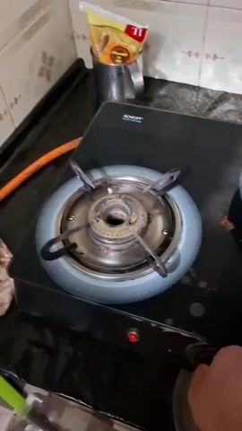 A person's hand, holding a lighter, approaches a gas stove. The lighter ignites, and a blue flame appears from the burner. The flame grows in intensity and then stabilizes, producing a consistent blue flame. Another burner on the stove is visible, but it is not in use. The background shows a kitchen counter with tiles and a container of cooking oil.